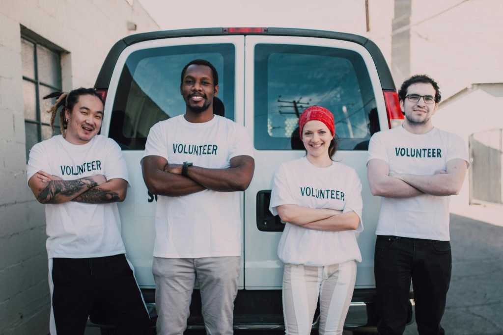 Diverse group of adult volunteers smiling in front of a van during a sunny day.